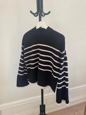 Svart stickad tröja med vita ränder - Säljer en svart stickad tröja med vita horisontella ränder. Tröjan har en hög hals, vida ärmar och ribbstickade detaljer vid hals, ärmslut och nederkant. Perfekt för dig som gillar en avslappnad och trendig look.