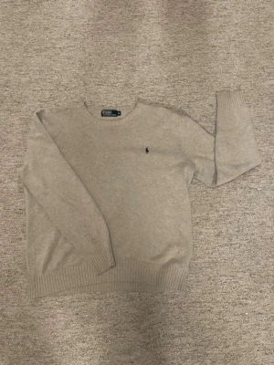 Beige stickad tröja från Polo Ralph Lauren - Snygg beige stickad tröja från Polo Ralph Lauren med klassisk rund hals och diskret logga broderad på bröstet. Tröjan har ribbade muddar vid ärmslut och nederkant, perfekt för lager-på-lager och chill stil.