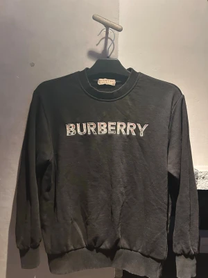 Svart sweatshirt från Burberry - Säljer en svart sweatshirt från Burberry med klassisk logotyp broderad i vitt och rött på bröstet. Tröjan har rund hals, ribbade muddar vid ärmslut och nederkant samt en avslappnad passform. Perfekt för en stilren och trendig look.