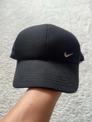 Svart Nike keps med silvrig logo - Snygg svart keps från Nike med klassisk silvrig swoosh-logga framtill. Kepsen har böjd skärm och är tillverkad i ett mjukt tygmaterial. Perfekt accessoar för en sportig och stilren look.