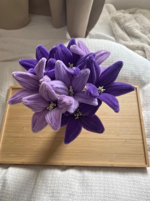 Bukett med lila tygblommor - En bukett med konstgjorda blommor i olika nyanser av lila och ljuslila. Blommorna är gjorda av mjukt tygmaterial och har gröna stjälkar. Perfekt som dekoration i hemmet och levereras i en enkel vit vas.