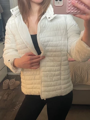 Herno liknande jacka - Säljer en stilren vit pufferjacka med hög krage och dragkedja framtill. Jackan har quiltad design och är lätt vadderad, perfekt för kalla dagar. Passar dig som vill ha en enkel men snygg vinterjacka. Jackan har en klassisk passform och är superenkel att matcha. Har ett par minimala fläckar, skriv för bild!