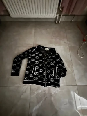 Svart kofta med vita GG-mönster - Snygg svart kofta  med vita GG-loggor över hela plagget. Koftan har dragkedja framtill, två fickor och ribbade muddar. Perfekt för dig som gillar exklusiva och trendiga plagg med tydlig logga.
