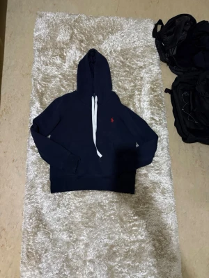 Mörkblå hoodie från Polo Ralph Lauren - Snygg mörkblå hoodie från Polo Ralph Lauren med klassisk röd logga broderad på bröstet. Hoodien har huva med vita dragsnören och en stor magficka. Perfekt för en avslappnad och stilren look.