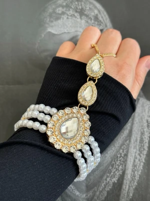 Guldigt armband med pärlor och stenar - Unikt armband med tre rader vita pärlor och stora droppformade glittriga stenar infattade i guldfärgad metall. Armbandet har en elegant design med gnistrande detaljer och passar perfekt för dig som vill sticka ut.