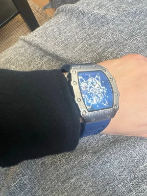 Richard Mille blå klocka - Säljer en Richard Mille klocka med blått armband och silverfärgat boett. Klockan har ett skeletturverk som syns genom den blå urtavlan och en robust, fyrkantig form. Armbandet är i syntetmaterial och boetten i metall. Perfekt för dig som gillar statement-accessoarer.