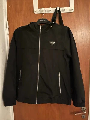 Svart vindjacka från Prada - Svart vindjacka från Prada med klassisk logotyp på bröstet. Jackan har silverfärgade dragkedjor, huva med dragsko och två fickor framtill. Materialet är glansigt och lätt, perfekt för att hålla vinden borta. Snygg och stilren design som passar till många outfits.