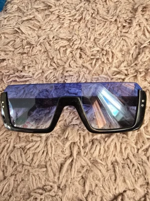 Svarta futuristiska solglasögon - Säljer ett par riktigt coola solglasögon med svart, bred båge och stora, blåtonade linser i futuristisk stil. Glasögonen har en rak, kantig form och små silvriga detaljer på sidorna. Perfekta för dig som vill sticka ut och addera edge till din look.