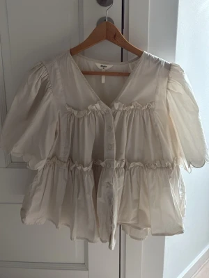 Vit volangblus med puffärm från Object - Supersöt vit blus från Object med volanger och knappar framtill. Blusen har puffiga korta ärmar och en luftig, flowy passform. Perfekt för en trendig och avslappnad look. Materialet känns lätt och svalt.