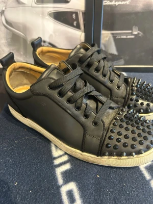 Svarta Louboutin sneakers med nitar - Snygga svarta sneakers från Christian Louboutin med ikoniska röda sulor och coola svarta nitar på tån. Skorna har svarta skosnören, låg profil och är tillverkade i slätt läder med beige innerfoder. Perfekta för dig som vill sticka ut med din stil.
