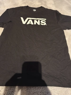 Svart T-shirt från Vans - Svart T-shirt från Vans med stort vitt logotryck på bröstet. Klassisk passform och rund halsringning. Perfekt för en avslappnad streetwear-look.