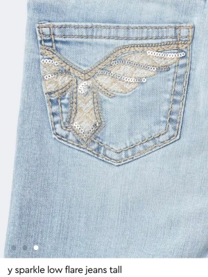 Ljusblå flare jeans med paljettdetalj - Säljer ett par ljusblå flare jeans med låg midja och slitningar på knäna. Bakfickan har en snygg broderad detalj med paljetter i silver och beige som ger extra bling. Perfekta för dig som gillar en retro och unik look.