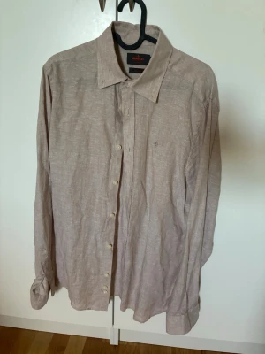 Beige linneskjorta från Morris - Snygg beige skjorta från Morris i 100% linne. Klassisk krage, knäppning framtill och lång ärm. Perfekt för en stilren och avslappnad look. Skjortan har en diskret logga broderad på bröstet och är i modellen Classic Fit.