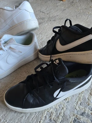 Nike sneakers svart och vit - Säljer ett par klassiska Nike sneakers i svart med vit sula och vit swoosh-logga på sidan. Skorna har lågt skaft och snörning framtill. Perfekta för en sportig och stilren look. Materialet är slätt läder och de har en tidlös design. 