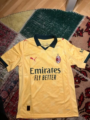 Gul AC Milan fotbollströja Puma - Snygg gul AC Milan matchtröja från Puma med svarta detaljer och klubbmärke på bröstet. Tröjan har korta ärmar, svart krage och tryckta loggor. Perfekt för fotbollsträning eller att bära som supporter. Materialet är lätt och andas bra.