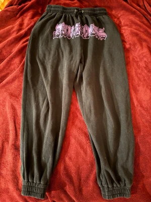 Svarta mjukisbyxor med lila tryck - Svarta mjukisbyxor med resår i midjan och benslut. Stort lila tryck med texten 'DARKNESS' framtill. Sköna och avslappnade med streetwear-vibe, perfekta för chill dagar. Materialet är mjukt och bekvämt, troligen bomullsblandning.
