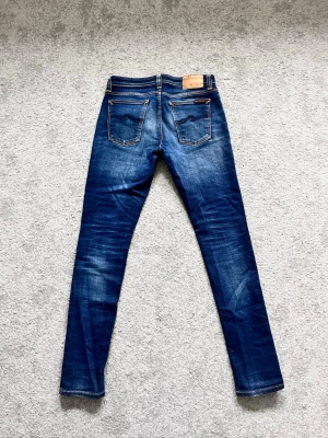 Blå slim fit jeans från Nudie Jeans - Säljer ett par blå slim fit jeans från Nudie Jeans med klassisk femficksdesign och snygga slitningar framtill. Jeansen har kontrastsömmar, läderpatch bak i midjan och är tillverkade i bomull med lite stretch för skön passform. Jeansen är lagade vid skrevet. Skriv vid minsta fundering!