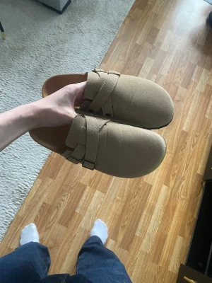 Beige tofflor - Säljer ett par snygga beige tofflor med justerbart spänne över foten. Ovandelen är i tyg och sulan är svart med mönstrat grepp. Perfekta för att glida runt hemma med stil.