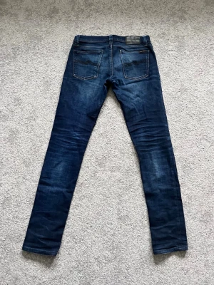 Mörkblå jeans från Nudie - Säljer ett par mörkblå jeans från Nudie Jeans med klassisk femficksmodell och snygga slitningar. Jeansen har kontrastsömmar och läderpatch bak i midjan. Perfekta för dig som gillar en smal passform och tidlös stil. Skriv vid minsta fundering! 