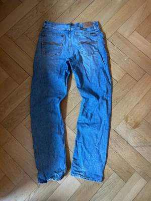 Blå jeans från Nudie Jeans - Klassiska blå jeans från Nudie Jeans med raka ben och femficksmodell. Jeansen har ljus tvätt och subtila slitningar, samt Nudie Jeans signatursöm på bakfickorna. Inga defekter utöver marginella slitningar i hälarna. Storlek 30/32. Nypris 1600. Tveka inte att höra av dig vid frågor eller funderingar.😊