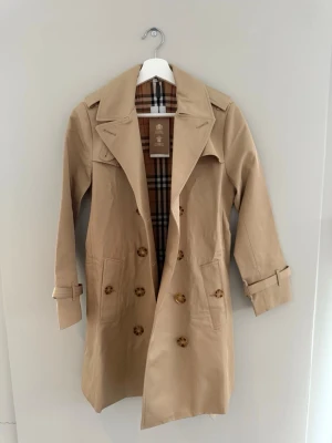 Beige trenchcoat från Burberry - Klassisk beige trenchcoat från Burberry med det ikoniska rutiga fodret. Jackan har dubbelknäppning, bred krage, bälte i midjan och justerbara ärmslut. Tillverkad i ett tåligt bomullsmaterial och har snygga detaljer som axelklaffar och stora knappar.