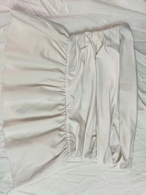 Vit kortkjol med volang - Trendig vit kortkjol med volangkant och rynkade detaljer i midjan. Kjolen har en axelbandslös look och är sydd i ett mjukt, lätt material som ger en luftig känsla. Perfekt för sommarens alla tillfällen.