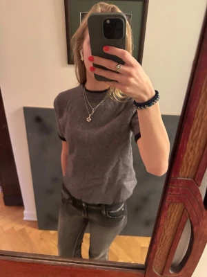 Grå stickad t-shirt med svarta detaljer - Snygg grå stickad t-shirt från Zara med svarta muddar vid ärmslut och halsringning. Tröjan har korta ärmar och en klassisk rund hals. Perfekt att styla med jeans eller kjol för en chill look.