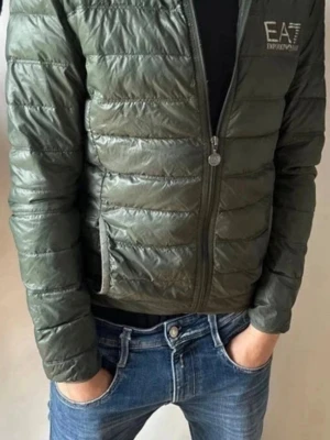 Grön dunjacka från EA7 Emporio Armani - Stilren grön dunjacka från EA7 Emporio Armani med quiltad design och diskret logga på bröstet. Jackan har dragkedja framtill, två sidofickor och lätt glansigt material. Perfekt för kalla vinterdagar och enkel att matcha till olika outfits. Storlek S