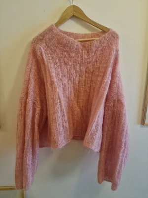 Rosa fluffig stickad tröja - Supermjuk och oversized handgjord stickad tröja i fluffigt rosa garn. Tröjan har v-ringning, långa ärmar och en lös passform som ger en chill vibe. Perfekt för lager-på-lager och att mysa i under kyliga dagar.