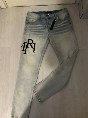 Ljusblå Amiri jeans - Snygga ljusblå skinny jeans med slitna detaljer och stora svarta bokstavstryck på både fram- och baksida. Jeansen har klassisk femficksdesign och är tillverkade i stretchigt denim för en bekväm passform. Perfekta för en trendig streetwear-look.