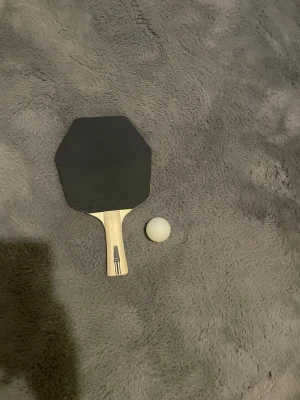 Pingisracket och boll i trä - Säljer ett pingisracket med ena sidan svart och andra sidan röd, samt en vit pingisboll. Racketen har ett handtag i ljust trä och en åttakantig form. Perfekt för att spela pingis med vänner eller familj. Obs går att upphämtas i södra Stockholm 