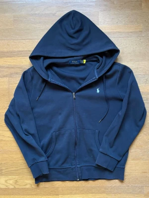 Marinblå hoodie från Polo Ralph Lauren - Snygg marinblå hoodie från Polo Ralph Lauren med dragkedja, huva och två fickor framtill. Klassisk broderad logga på bröstet och ribbade muddar. Perfekt för en chill och stilren look. Materialet är mjuk bomullsblandning.