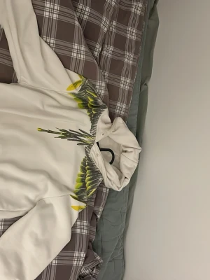 Marcelo burken tröja - Säljer en vit hoodie med unikt tryck av gröna och gula fjädrar över axlar och bröst. Hoodien har huva och en avslappnad passform, perfekt för dig som vill sticka ut med en cool design. Materialet känns mjukt och bekvämt mot huden.