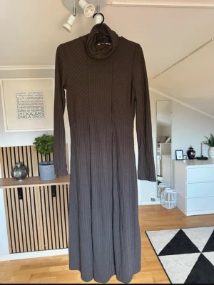 Brun ribbad långklänning med polokrage - Säljer en brun långklänning med ribbad struktur och polokrage. Klänningen har långa ärmar och en figurnära passform som ger en snygg siluett. Perfekt för dig som gillar stilrena och bekväma plagg med en twist.