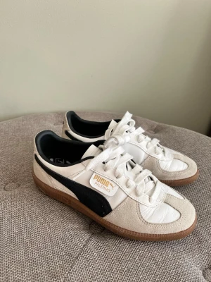 Puma Palermo vita sneakers - Snygga Puma Palermo sneakers i vitt läder med beige mockadetaljer och klassisk svart Puma-stripe på sidan. Gummisula i brunt och vita skosnören. Märket syns tydligt på sidan och sulan. Perfekta för dig som gillar retrostil och streetwear. Använda 2-3 gånger. 