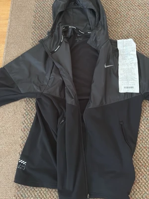 Svart Nike vindjacka med huva - Snygg svart vindjacka från Nike med huva och dragkedja. Jackan har vita Nike-loggor, sidofickor med dragkedja och detaljer med texten 'NIKE RUNNING' på insidan av luvan. KVITTO FINNS MED KÖPT FÖR 999 kr för 1 vecka sedan