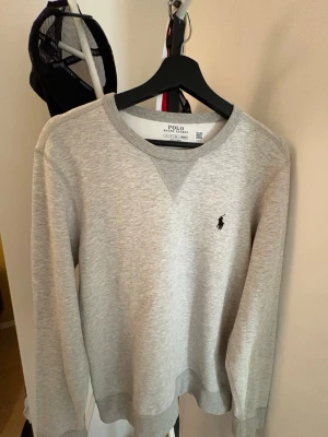 Grå sweatshirt från Polo Ralph Lauren - En aldrig använd snygg ljusgrå sweatshirt från Polo Ralph Lauren med klassisk rund halsringning och den ikoniska lilla loggan broderad på bröstet. Tröjan har ribbade muddar vid ärmslut och nederkant. Perfekt för en clean och avslappnad stil.