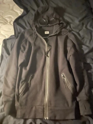 Svart hoodie med glasögon från C.P. Company - Svart hoodie från C.P. Company med dragkedja framtill och två fickor med dragkedja. Huvan har inbyggda glasögon som ger en unik och cool look. Tillverkad i 100% bomull för skön komfort. Perfekt för dig som vill sticka ut med streetwear-vibe.