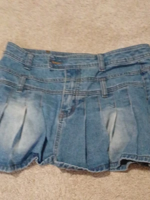 Blå jeansskort med veck - Snygg blå jeansskort med markerade veck framtill och baktill. Klassisk denimlook med bälteshällor och ljusa slitningar för en trendig känsla. Perfekt för dig som gillar en avslappnad men ändå stilsäker vibe.