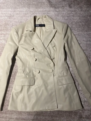 Beige kavaj i skinnimitation från ZARA - Stilren beige kavaj från ZARA i skinnimitation med dubbelknäppning och markerad midja. Kavajen har breda slag, två fickor med lock och en snygg, figurnära passform. Öppen för prisändring.