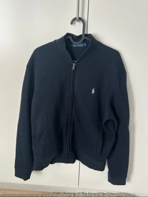 Svart zip-hoodie från Polo Ralph Lauren - Klassisk svart hoodie från Polo Ralph Lauren med dragkedja framtill och broderad logga på bröstet. Tröjan har ribbade muddar och två fickor. Perfekt för en avslappnad och stilren look. Storlek S pris kan diskuteras 