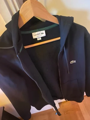  Lacoste Tröja - Svart hoodie med dragkedja från Lacoste, klassisk passform. Har huva med dragsko, två fickor framtill och den ikoniska gröna krokodilloggan på bröstet. Perfekt för en chill och stilren look.