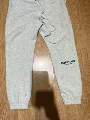 Grå Essentials mjukisbyxor - Snygga ljusgrå mjukisbyxor från Fear of God Essentials med svart logga tryckt på benet. Byxorna har dragsko i midjan och resår vid bensluten för en skön passform. Perfekta för en avslappnad och trendig stil.