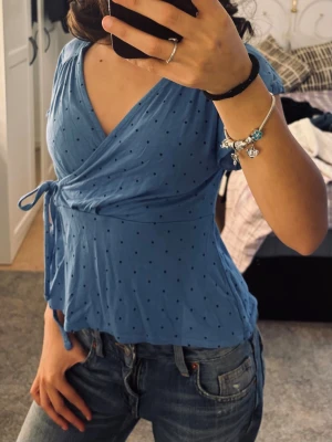Blå omlottopp med prickigt mönster - Snygg blå omlottopp med små svarta prickar och knyt i sidan. Toppen har korta ärmar och v-ringad halsringning, vilket ger en feminin och trendig look. Perfekt att matcha med jeans för en avslappnad stil.