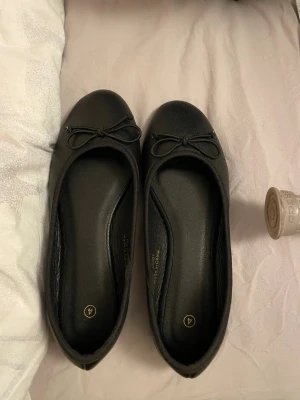 Svarta ballerinaskor med rosett - Säljer ett par klassiska svarta ballerinaskor med rund tå och söt rosettdetalj framtill. Skorna har platt sula och är tillverkade i syntetmaterial med en mjuk finish. Perfekta för dig som gillar enkel och stilren design. Aldrig använda. De är i storlek 37