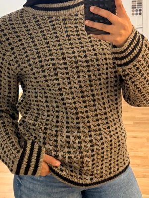 Beige och svart stickad tröja i ull - Stickad tröja från i 100% ull med beige botten och svart mönster. Tröjan har rund hals, långa ärmar och ribbade muddar med svarta ränder. Perfekt för dig som gillar klassisk men ändå unik stil. Ej använd. Prislappen fortfarande på 