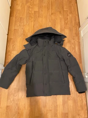 Svart dunjacka från Canada Goose - Säljer en svart dunjacka från Canada Goose med huva och hög krage. Jackan har två stora framfickor med dragkedja och tryckknappar framtill. Klassisk patch-logga på armen och en stilren, rak .