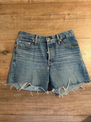 Levi's blå jeansshorts med rå kant - Klassiska blå jeansshorts från Levi's med hög midja och rå, fransig kant. Shortsen har fem fickor, bälteshällor och knappgylf. Tillverkade i slitstark bomull med den ikoniska Levi's-lappen bak. Perfekta för sommaren!