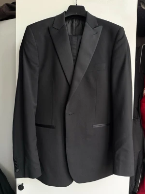 Smoking från Royal Black - Stilren svart smoking från Royal Black med klassiska slag och diskreta fickor. Kavajen har en elegant finish och är helfodrad, perfekt för dig som vill ha en tidlös look. Materialet är troligtvis polyester eller ullblandning. 46 i byxor, 146 i kavaj.