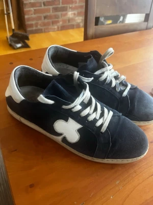 Marinblå sneakers från The Gallerist - Snygga marinblå sneakers med vit klöverdetalj på sidan och vita partier vid häl och snörning. Skorna har vita snören och en låg silhuett. Ovandelen verkar vara i mocka eller liknande material. Perfekta för dig som gillar unik design och streetstyle.